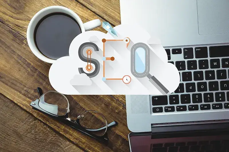 Laptop auf einem Tisch daneben eine Tasse Kaffee und als Overlay eine Cloud-Wolke mit dem Wort SEO darin